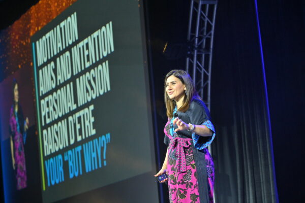 Shabnam Mogharabi stands on stage wearing a navy and pink floral dress. The slide in the background says "Motivational goals and intention personal mission reason for your “But why?” »"