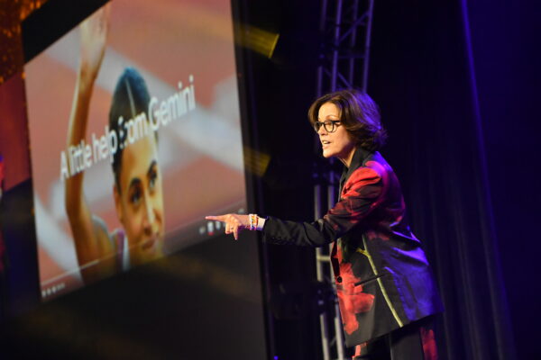 Ann Handley wears black glasses and a metallic burgundy suit pointing at the audience. The slide in the background says "A little help from Gemini."