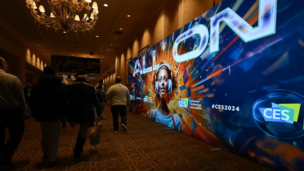 CES 2024 attendees walk past a large digital billboard that reads "EVERYTHING" on a photo of a woman with headphones. Explosion of fire behind his head.