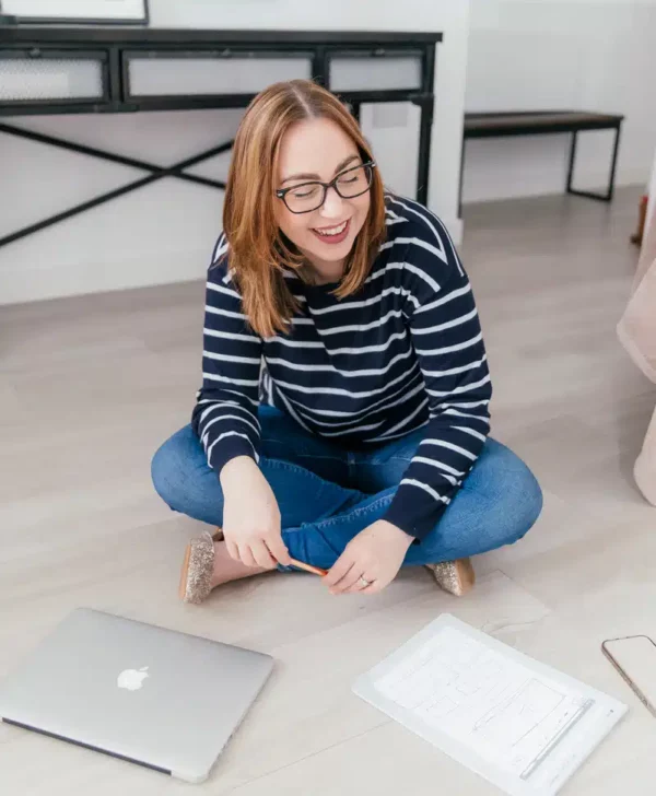 Kate is sitting on the floor, wearing a blue and white sweater and blue jeans. She wears glasses and looks down smiling.